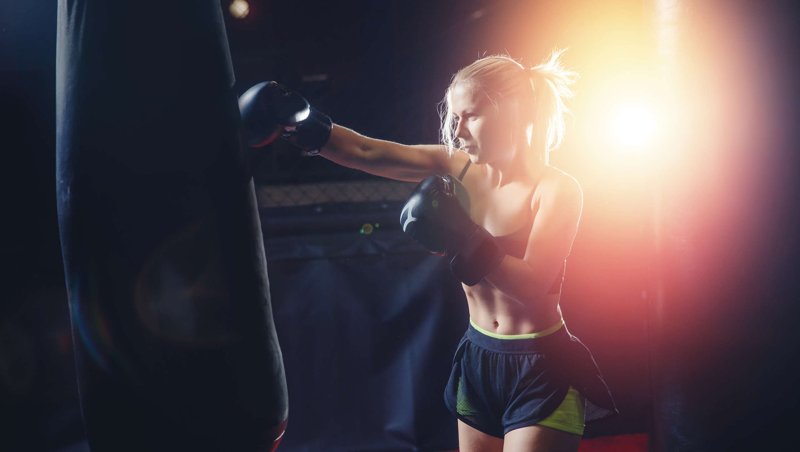 Female boxer MMA in sports shorts and short crop top trains gym, works out right punch on punching bag, preparing for battle. Dark background, bright red glare from flash